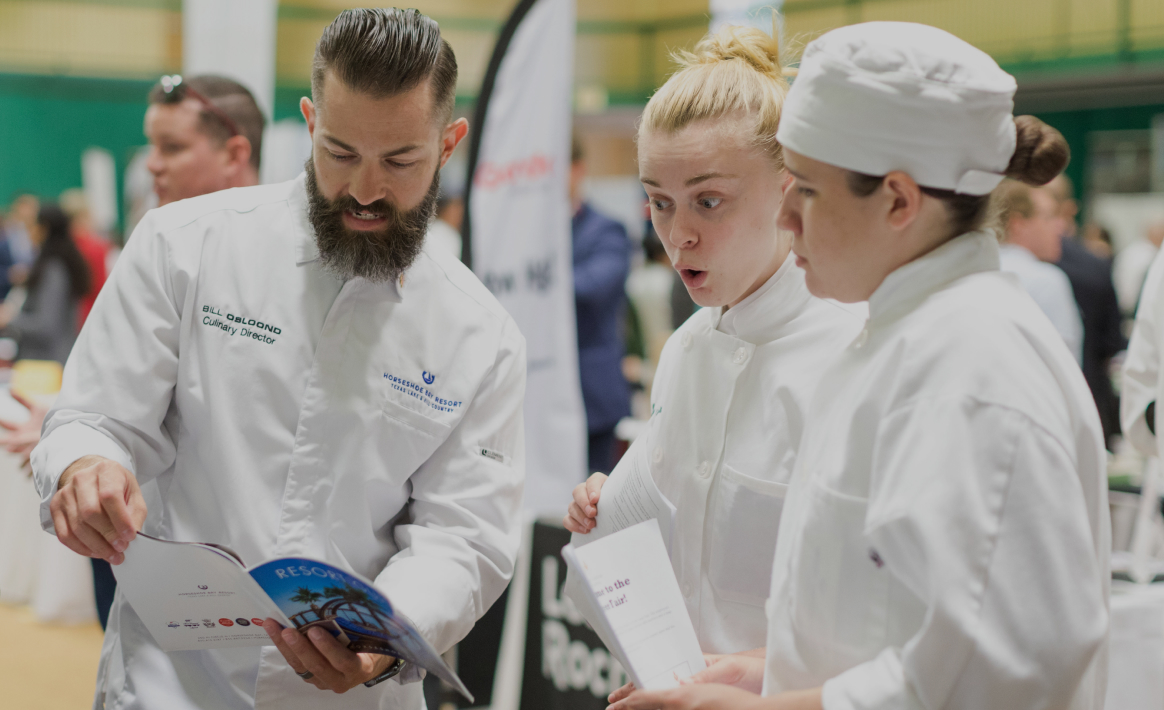 Three chefs, two women and one man, are wearing white uniforms and engaged in an attentive discussion about food service management. The man is holding and pointing to an open magazine while the others look on with interest. The background shows a blurred indoor event setting.