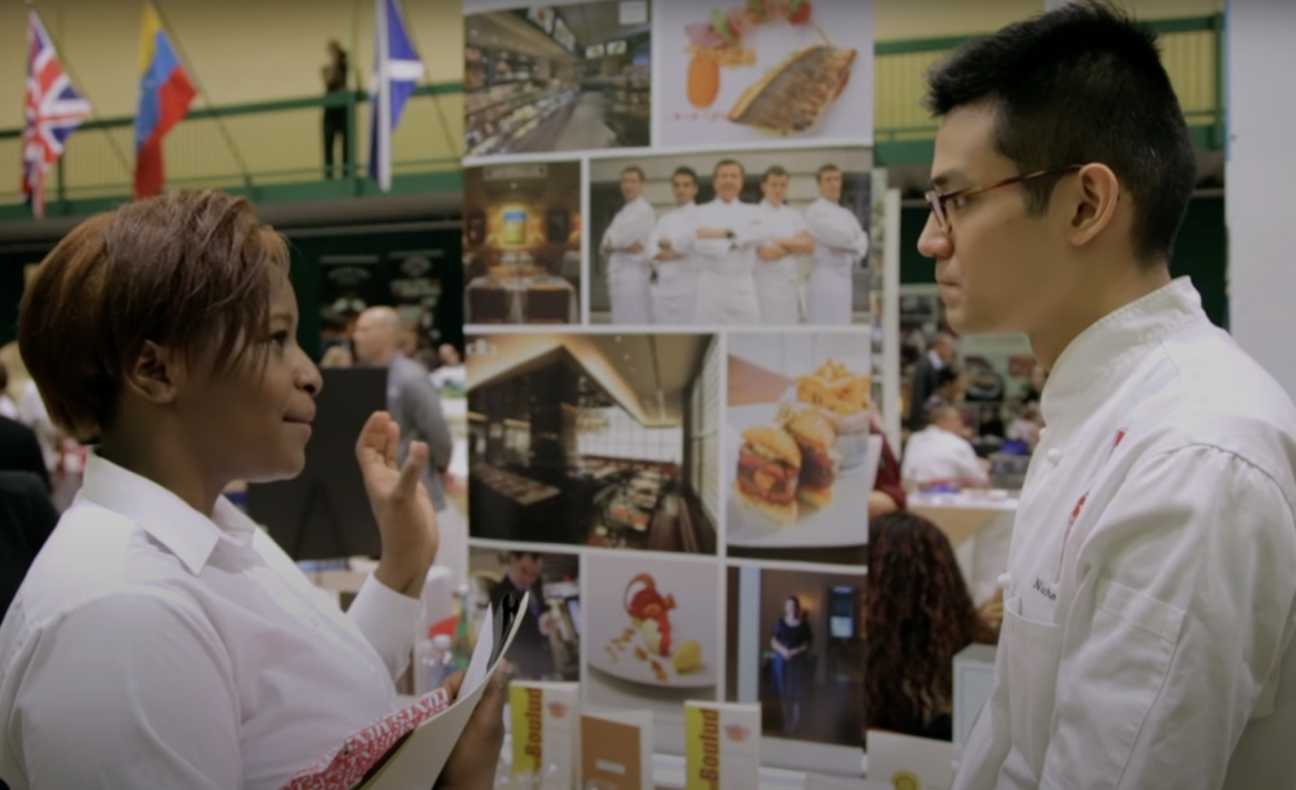 Two people engaged in conversation at an event. The woman on the left is gesturing while holding a notebook and pen, discussing aspects of food service management. The man on the right, wearing a chef's uniform, is listening intently. Behind them, large posters feature images of food and chefs. National flags hang above.