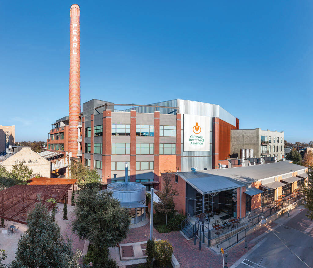 A panoramic view of a large brick building with a tall smokestack labeled "PEARL." The building houses the Culinary Institute of America, as indicated by a prominent sign on the facade. Surrounding the building are trees, pathways, and adjacent structures.