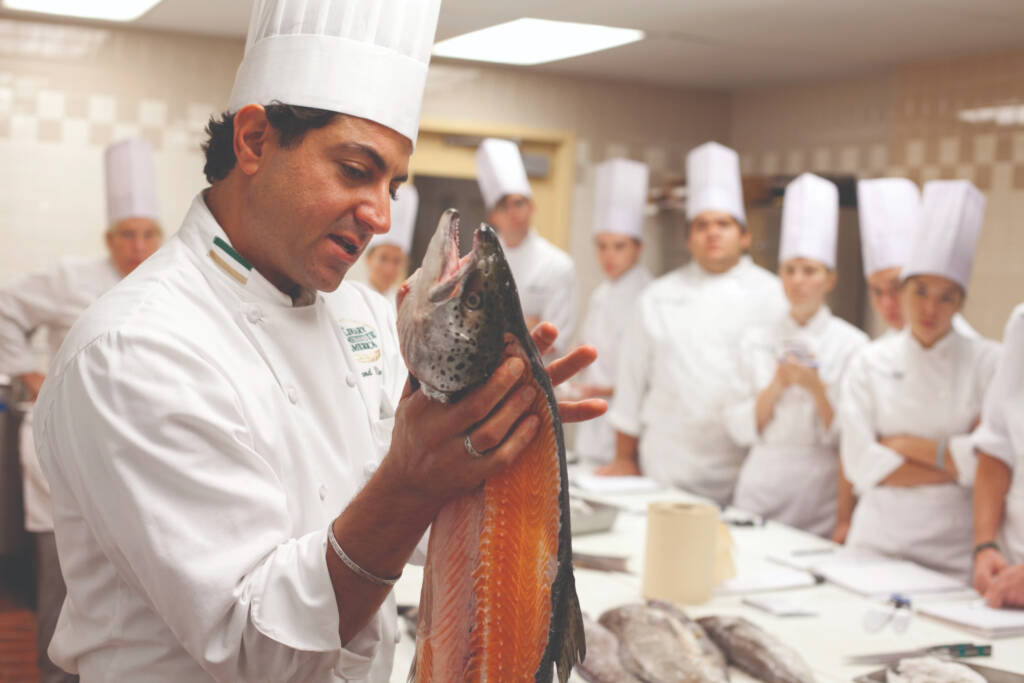 A chef in a white uniform and tall white hat holds up a large fish and appears to be explaining something to a group of students, who are also dressed in white chef uniforms with tall hats, in a professional kitchen setting.