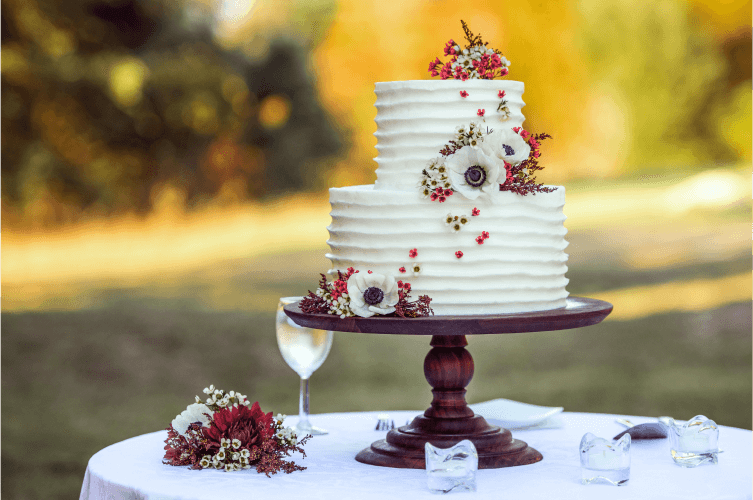 A two-tiered white wedding cake decorated with flowers and berries sits on a wooden stand outdoors. The cake has a textured design, and the table also holds a wine glass, floral arrangement, and small candles, with a blurred natural background.