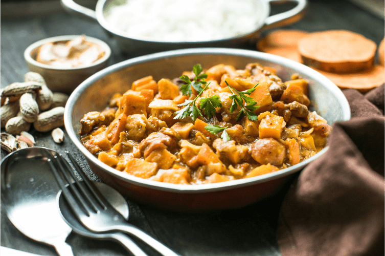 A bowl of peanut chicken stew garnished with fresh parsley sits on a rustic table. Silverware rests nearby, along with a napkin. Surrounding ingredients include whole peanuts, a small bowl of peanut butter, and sliced fresh vegetables.