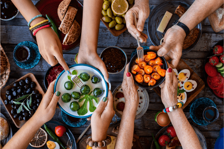 Four hands with forks reaching for plates of vibrant, healthy food, including Brussels sprouts and roasted vegetables, on a wooden table laden with a variety of dishes such as cheese, fruit, and deviled eggs. This scene at a culinary school for adults suggests a convivial meal gathering.