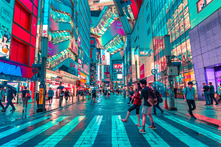 A vibrant, neon-lit city street at night with numerous billboards and signs. People walk across a crosswalk, some holding umbrellas. The buildings are lined with colorful advertisements and lights, creating a busy, lively atmosphere.