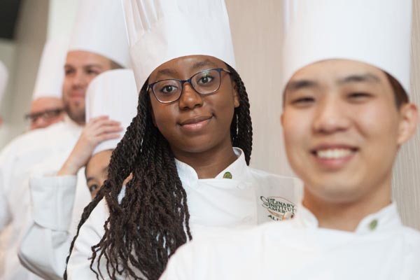 A group of chefs in white uniforms and tall white hats stand in a line, smiling at the camera. The front chef is an Asian man, followed by an African American woman with long braids and glasses. Other chefs can be seen in the background.