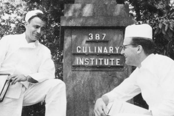 Black-and-white photo of two chefs in white uniforms and hats standing by a stone sign that says "387 Culinary Institute." One chef is leaning on the sign, and the other is kneeling beside it, both smiling at each other. Trees are visible in the background.