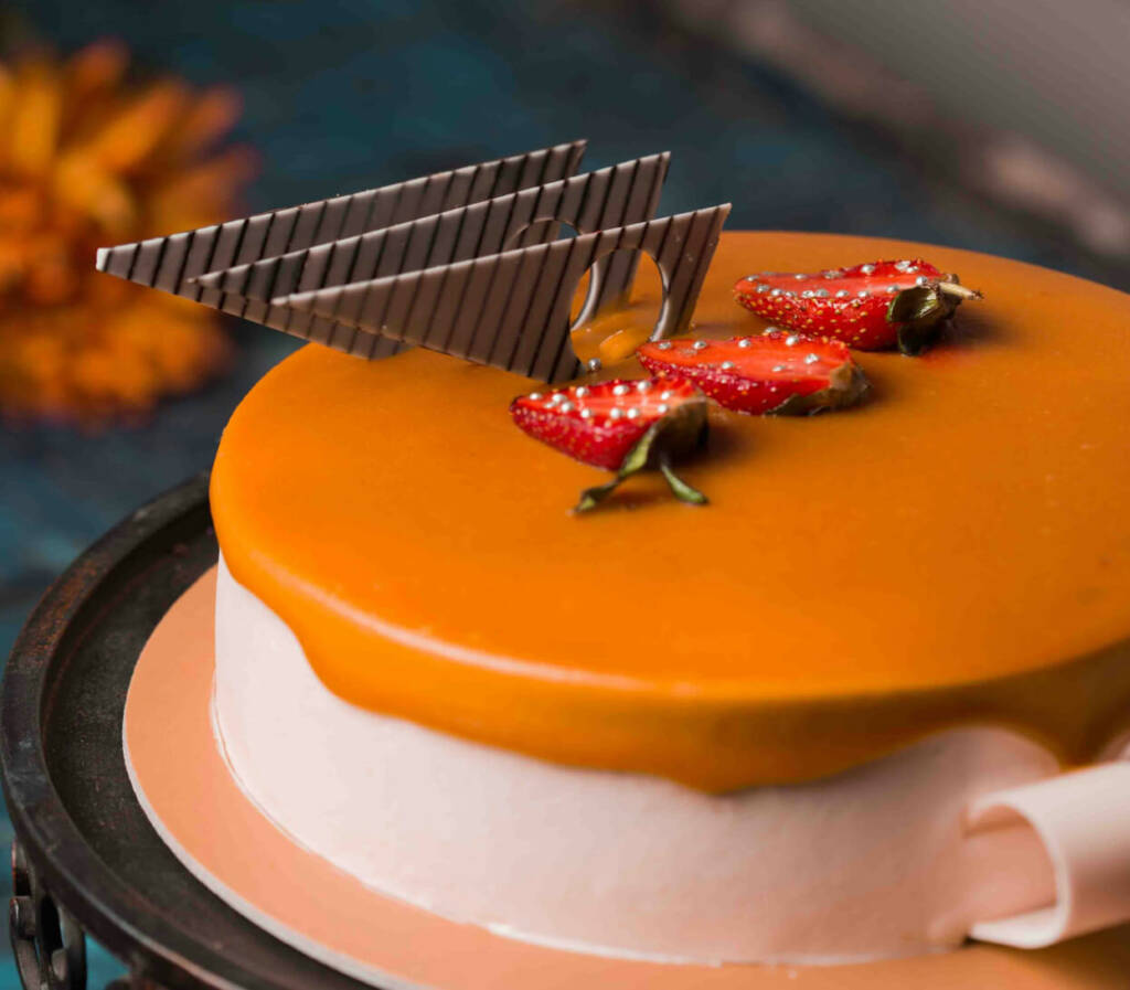 A round, smooth cake with a glossy caramel-colored topping and a white frosting base. The cake is garnished with fresh strawberry halves and a decorative piece of striped chocolate. A blurred flower can be seen in the background.