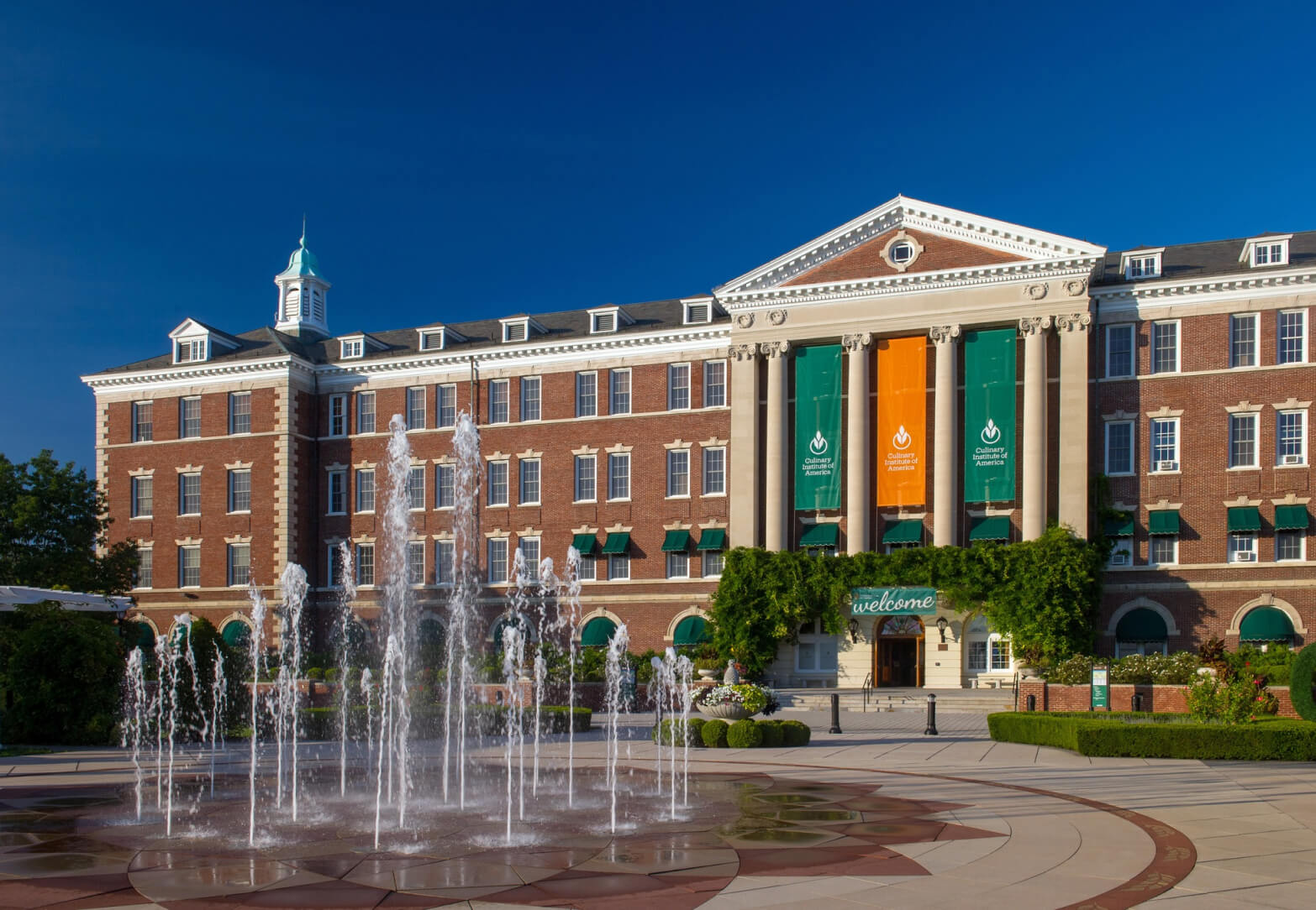 A large, stately brick building with a white facade and green awnings, featuring a prominent "Welcome" sign and banners. In front, a circular plaza with an elegant fountain sprays water into the air, surrounded by well-manicured greenery under a clear blue sky. It resembles one of the distinguished Culinary Institute of America locations.