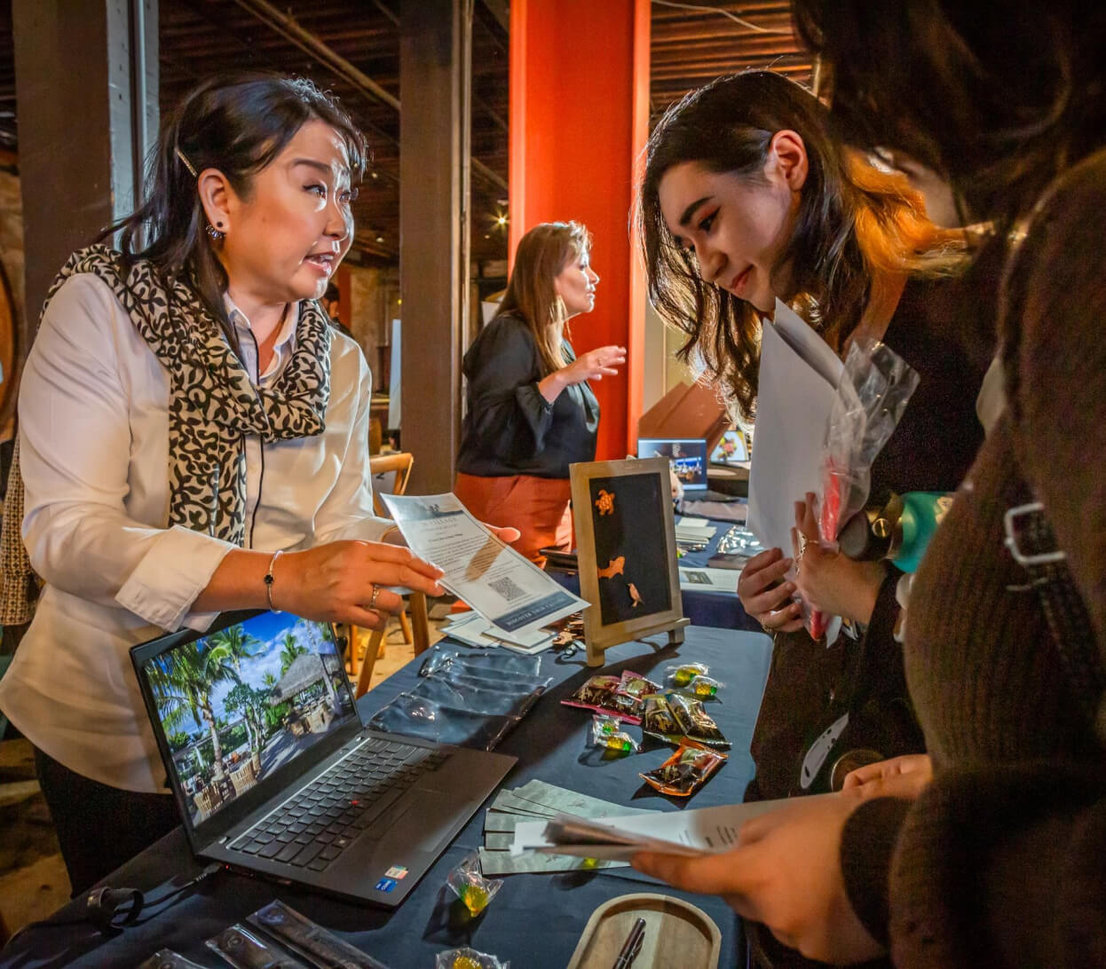 A woman at a booth is presenting information to two individuals at an indoor event. There is a laptop on the table displaying an image. Other items and pamphlets are spread out on the table. Several people are visible in the background, engaging in conversations.