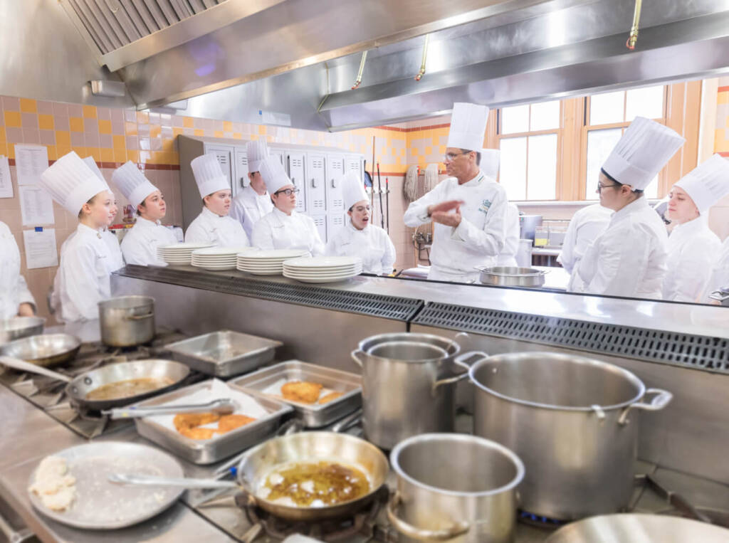 A chef in a white uniform and hat instructs a group of eight students, also in white uniforms and hats, in a commercial kitchen. The kitchen features several pots, pans, and stacks of plates. Stainless steel appliances and a tiled wall are visible in the background.