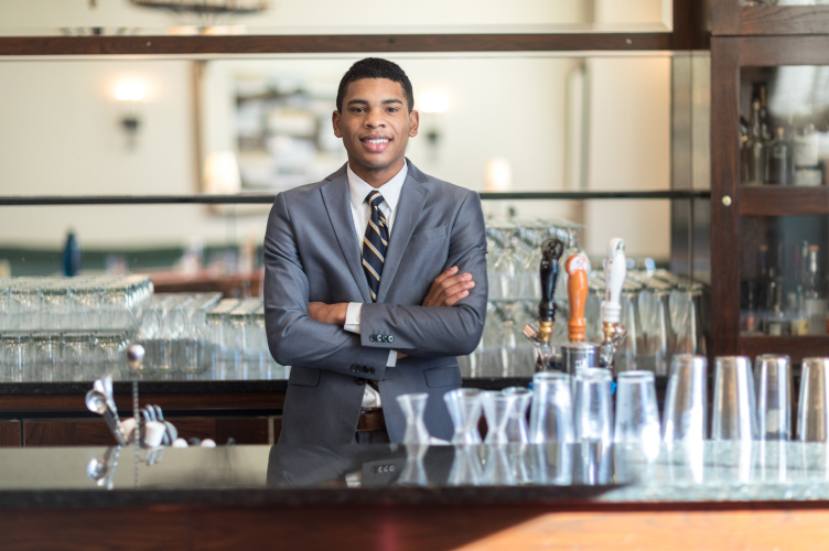 A man in a gray suit with a striped tie stands behind a bar, smiling with his arms crossed. Several glasses, taps, and bar tools are visible on the counter in front of him. The background features shelves with various bottles and glasses.