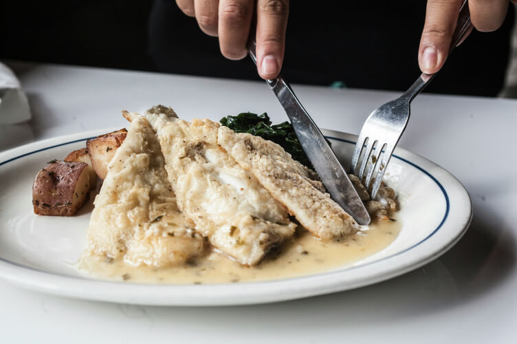 A person uses a knife and fork to cut into a piece of fish covered in creamy sauce on a white plate. The dish also includes roasted red potatoes and a serving of sautéed greens.