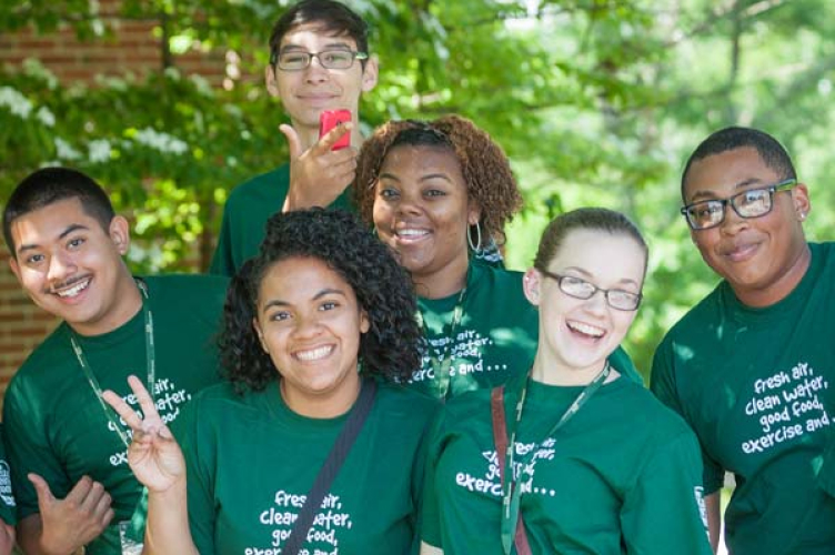 A group of six diverse young people wearing matching green t-shirts smiles at the camera. They are outdoors with trees in the background. One person holds up a peace sign, and another holds a red phone. The t-shirts say, "Fresh air, clean water, good food, exercise", promoting CIA admissions.