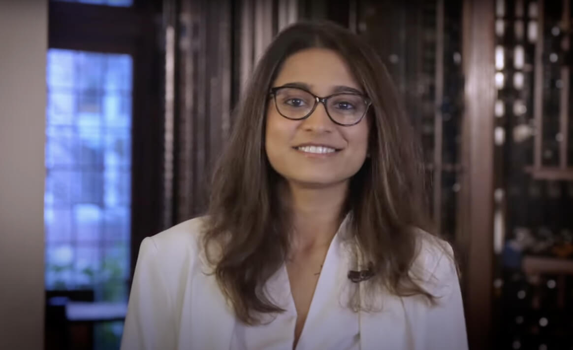 A woman with long brown hair and glasses is smiling at the camera. She is wearing a white blazer and standing indoors, with a background featuring large windows and some blurred objects.