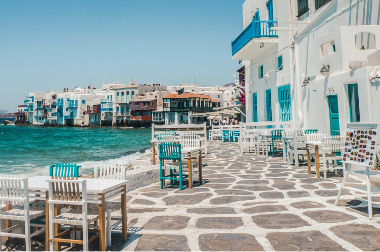 Oceanfront dining area with white wooden tables and chairs on a stone-paved walkway. White buildings with colorful balconies line the background, overlooking the turquoise sea on a sunny day. The scene exudes a laid-back coastal charm.