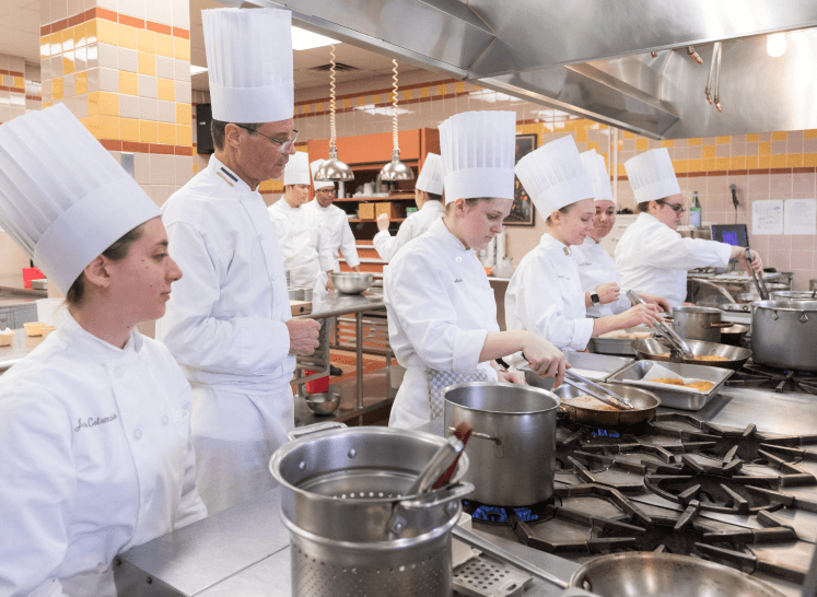 A group of chefs and culinary students in white uniforms and tall chef hats are cooking in a professional kitchen at the CIA Hyde Park Campus. They are busy at various stovetops, stirring pots and preparing food. The kitchen is equipped with large pots, pans, and industrial appliances.