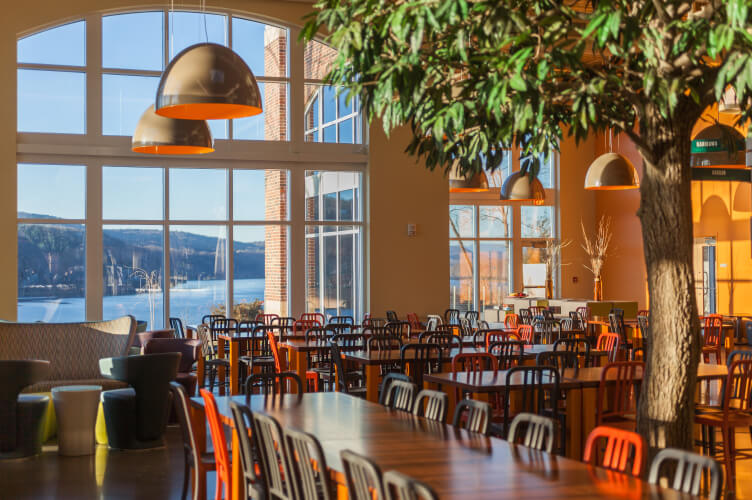 A spacious cafeteria on the CIA Hyde Park campus features numerous tables and chairs, large windows offering a scenic view of a lake with mountains in the background. Warm sunlight streams in, illuminating the modern décor and vibrant orange light fixtures hanging from the ceiling.