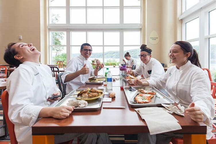 A group of four people wearing chef uniforms are sitting at a table in a brightly lit dining area on the CIA Hyde Park campus, smiling and enjoying their meal. One person is laughing heartily, while the others appear to be engaged in conversation and eating. Trays of food are laid out on the table.