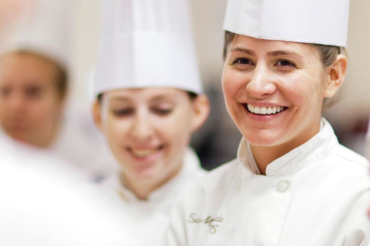 Two smiling chefs in white uniforms and tall white chef hats. The focus is on the chef in the foreground, who has a white jacket with indistinct text on it, while the chef in the background is slightly out of focus. Both appear to be in a kitchen setting.