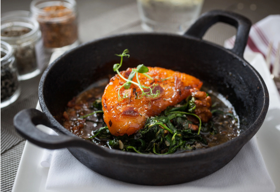 A cast iron skillet holds a cooked piece of glazed salmon garnished with microgreens, served over a bed of sautéed leafy greens. The dish is presented on a white napkin with jars of spices and a glass of water blurred in the background.