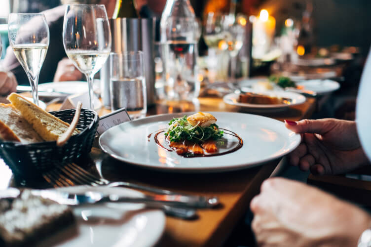 A close-up view of a gourmet meal with various dishes on a wooden table in a restaurant setting. The main focus is a plated dish with garnish, surrounded by wine glasses, cutlery, and a bread basket. Soft, warm lighting enhances the cozy ambiance.