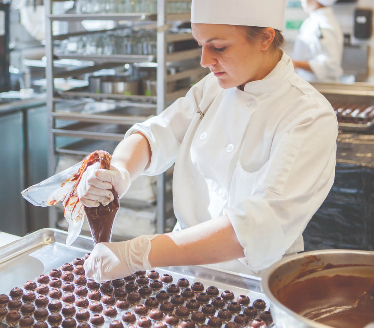 A pastry chef in a white uniform and hat is piping chocolate into molds in a commercial kitchen. She is focused on her task, handling a piping bag filled with chocolate mixture, with trays and cooking equipment visible in the background.