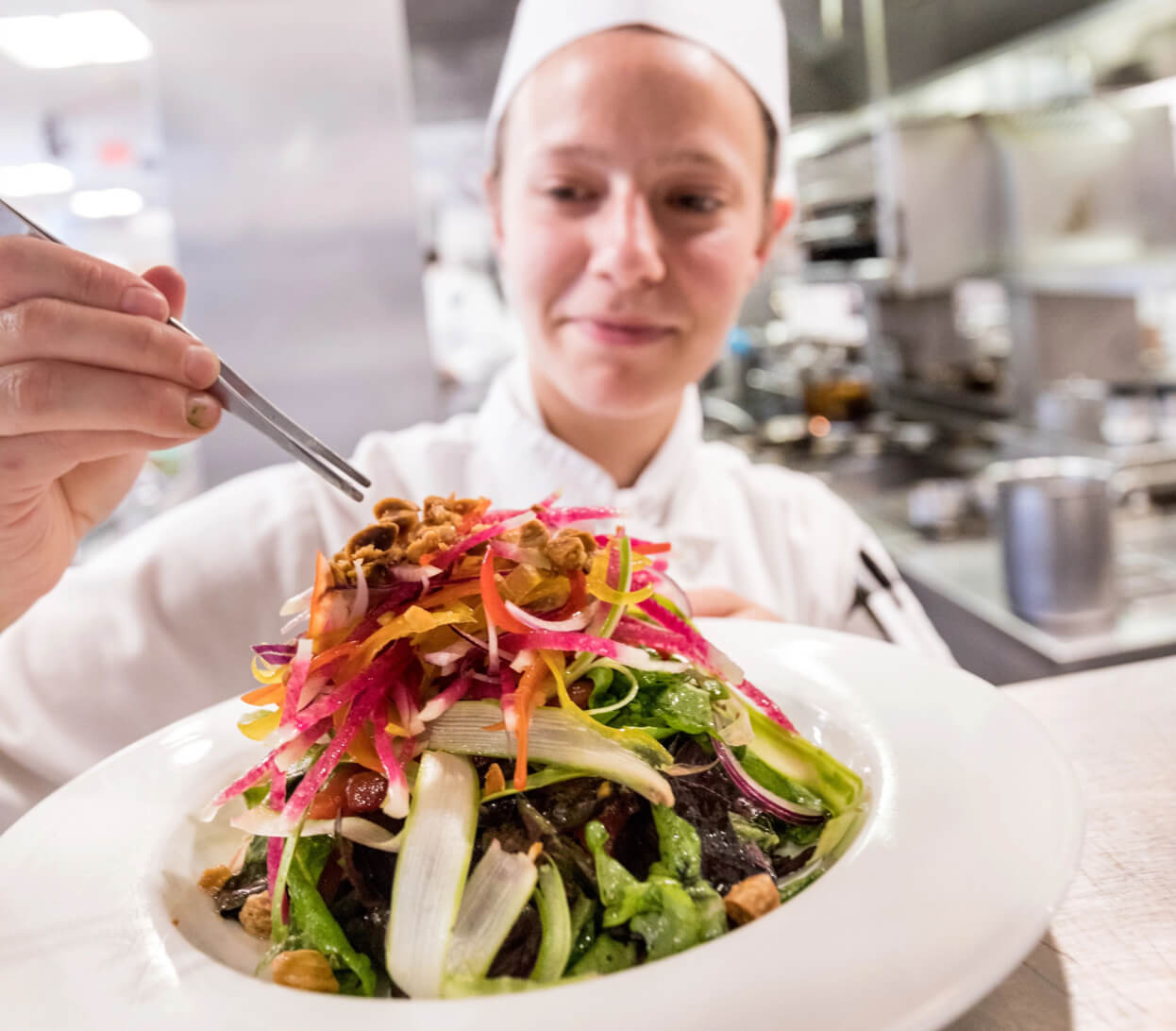 A chef in a white uniform and hat is carefully garnishing a colorful salad with tweezers in a professional kitchen. The salad includes vibrant greens, sliced vegetables, and what appears to be nuts. The kitchen background is blurred, focusing attention on the chef and the dish.