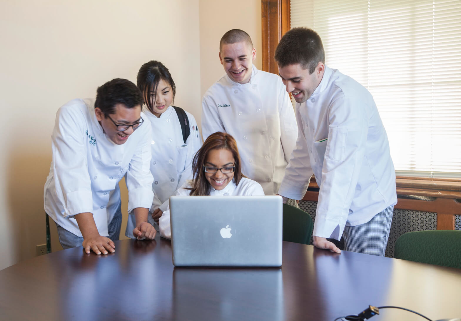 Four chefs in white uniforms gather around a laptop, smiling and looking at the screen. They are standing in a well-lit room with a large window. The individual in the middle is seated, while the others stand around the table, discussing culinary school financial aid options.