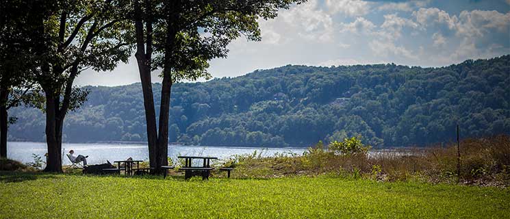 A person sits in a chair under the shade of tall trees, overlooking a serene lake surrounded by lush, green hills. A picnic table is nearby, and the sky is partly cloudy with ample sunlight. The scene evokes a sense of calm and relaxation.