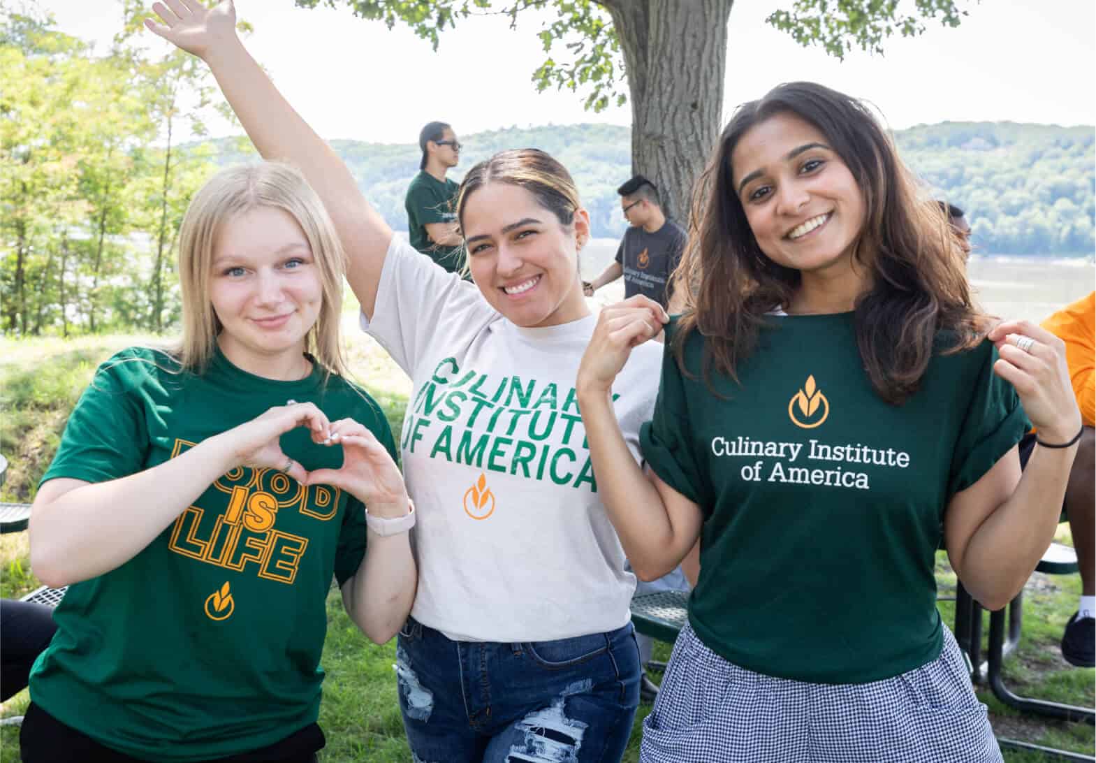 Three smiling women, likely admitted students, are posing outdoors, wearing t-shirts from the Culinary Institute of America. One makes a heart symbol with her hands. A lake and trees are in the background. Other people are seen mingling in the distance.