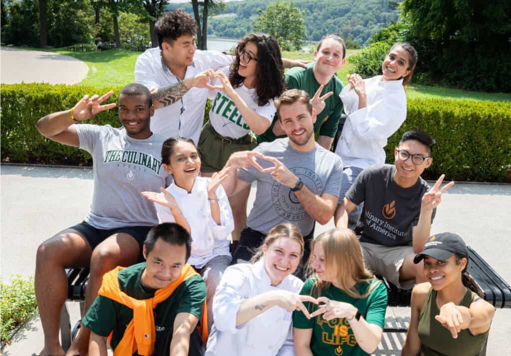 A group of young adults, some in culinary uniforms, pose outdoors with playful smiles and hand signs. They are seated on a bench and standing around it, with a lush green landscape in the background. The admitted students create a cheerful and friendly atmosphere.