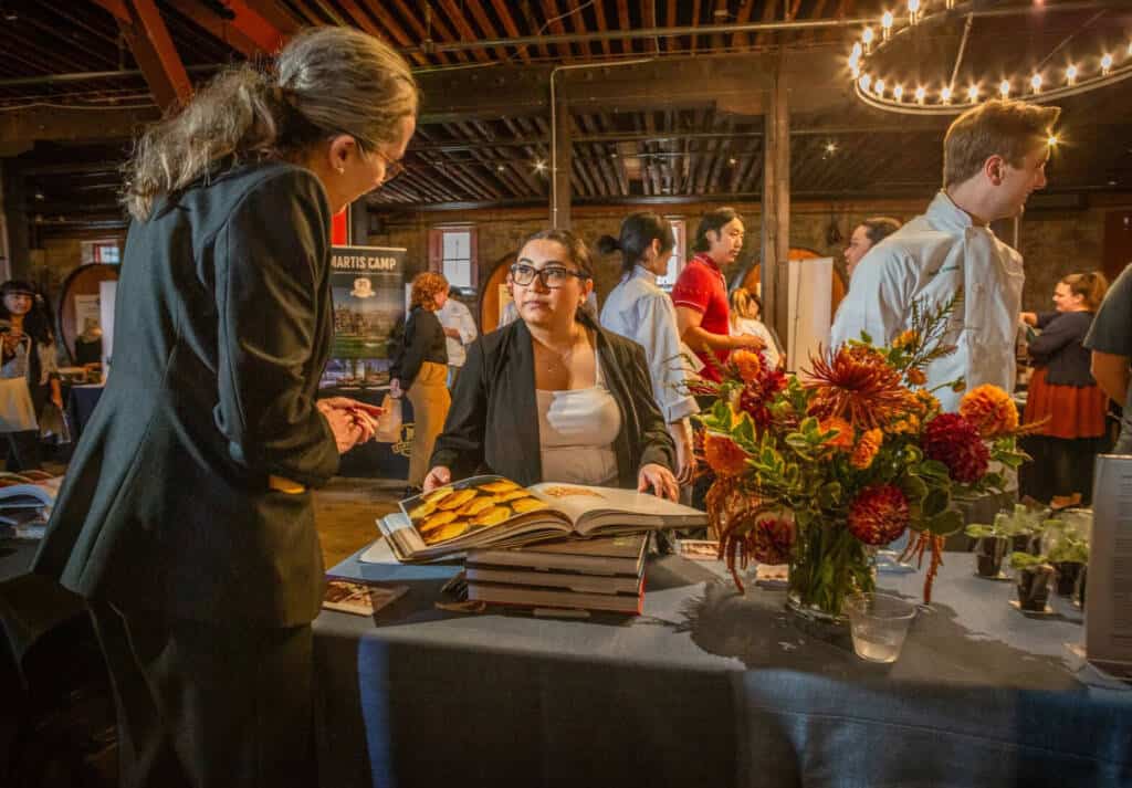 A woman with glasses sits behind a table displaying an open book and vibrant flowers in a warmly lit room. She is engaged in conversation with another woman standing across from her. Various booths and attendees are visible in the background.