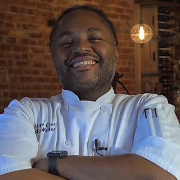 A smiling chef with a beard, dressed in a white chef's jacket, stands with arms crossed in front of a rustic brick wall. A wooden shelf with various items is visible in the background, along with warm, ambient lighting.