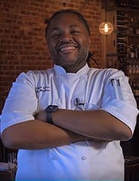 A chef wearing a white uniform stands confidently with arms crossed in a warmly lit restaurant. There is a brick wall and wine bottles visible in the background. The chef is smiling broadly and has a beard.