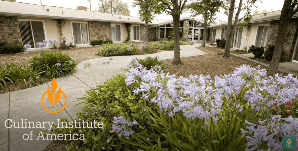 A building complex with a garden filled with purple flowers and green plants in the foreground. Trees and shrubs are scattered around the courtyard. The text "Culinary Institute of America" with an orange flame logo is overlaid on the image in the lower left corner.