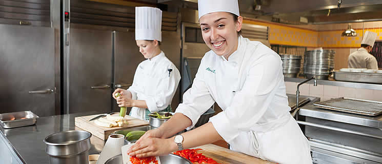 Two chefs in professional kitchen attire are preparing food. The chef in the foreground smiles while cutting red bell peppers, and the chef in the background is focused on peeling potatoes. Stainless steel appliances and utensils are visible in the kitchen.