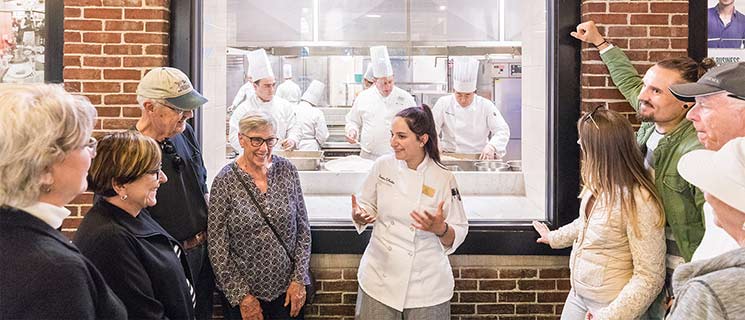 A chef in a white coat is speaking to a group of attentive people about culinary programs in front of a large window into a kitchen. Inside the kitchen, multiple chefs in white uniforms and hats are working. The group stands in front of a brick wall with the kitchen window behind them.