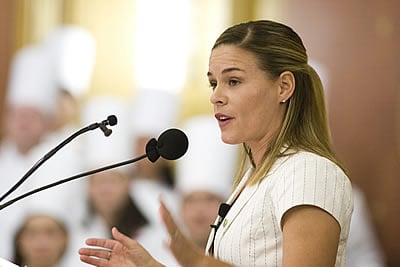 A woman with light brown hair, tied back, is speaking into a microphone. She is wearing a white top and is positioned slightly to the right. In the background, several people wearing white chef hats are visible, though they are out of focus.