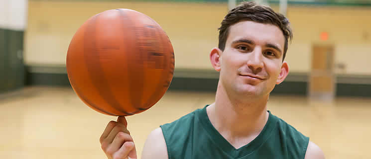 A young man in a green basketball jersey is in a gymnasium, balancing a basketball on the tip of his index finger. He has short dark hair and is smiling slightly at the camera. The background features a blurry gym setting with a door and basketball court markings.