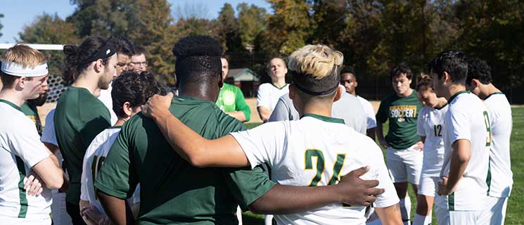 A group of soccer players stand in a circle on a field, wearing green and white uniforms. Some players have their arms around each other's shoulders, appearing to listen to a person in the center. Trees with autumn foliage are in the background.