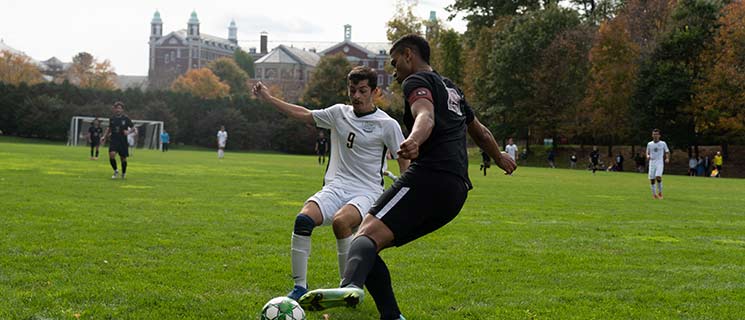 Two soccer players vie for control of the ball on a grassy field. The player in white, wearing number 9, challenges the player in black, wearing number 22, who is kicking the ball. Other players and a building are visible in the background. The scene is set during the day.