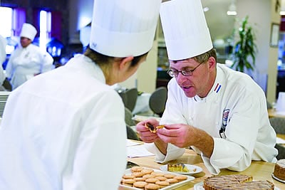 Two chefs wearing white uniforms and tall chef hats are working together in a kitchen. One chef is holding a small item, possibly food, and explaining something to the other chef. A table in front of them has plates with cookies and other food items.