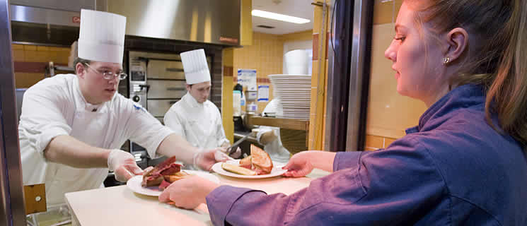 A chef in a white uniform and tall hat hands a plate of food through a serving window to a woman in a blue shirt, who dreams of applying for grants for culinary school. Another chef in the background holds a plate, showcasing the bustling atmosphere of the commercial kitchen setting.