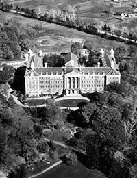 Aerial black and white photo of a large, historic building with tall columns at the entrance, surrounded by trees and expansive lawns. The structure has a symmetrical design with multiple stories and towers, set in a rural landscape.