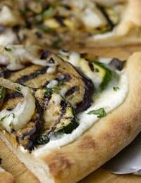 A close-up of a slice of pizza topped with grilled zucchini, onions, and melted cheese on a light, fluffy crust. The toppings are slightly charred, giving a rustic appearance to the delicious-looking slice.