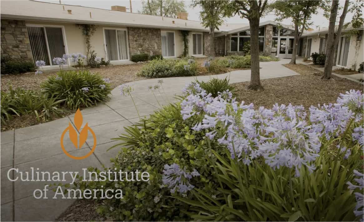 The image shows an exterior view of the Culinary Institute of America building. The pathway is lined with blooming purple flowers and lush greenery, leading to the entrance. The institute's logo and name are displayed in the lower-left corner.