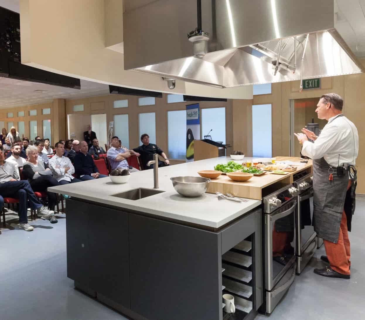 A chef in an apron gives a cooking demonstration in a kitchen setup with stainless steel appliances and countertops, facing an audience seated in rows. The audience is attentively watching, and a banner is visible in the background.