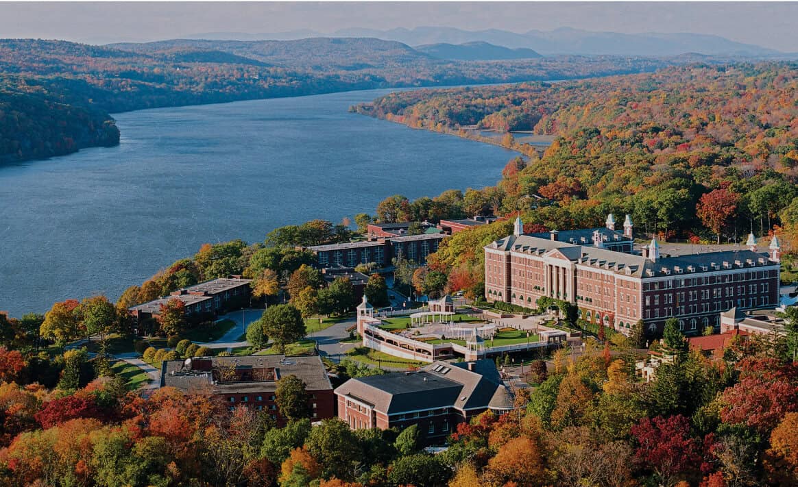 Aerial view of a campus situated in a lush area with vibrant autumn foliage. The Culinary Institute of America, featuring large brick buildings with white accents, is nestled along a serene river that winds through the landscape. Rolling hills covered in fall trees extend into the distance.