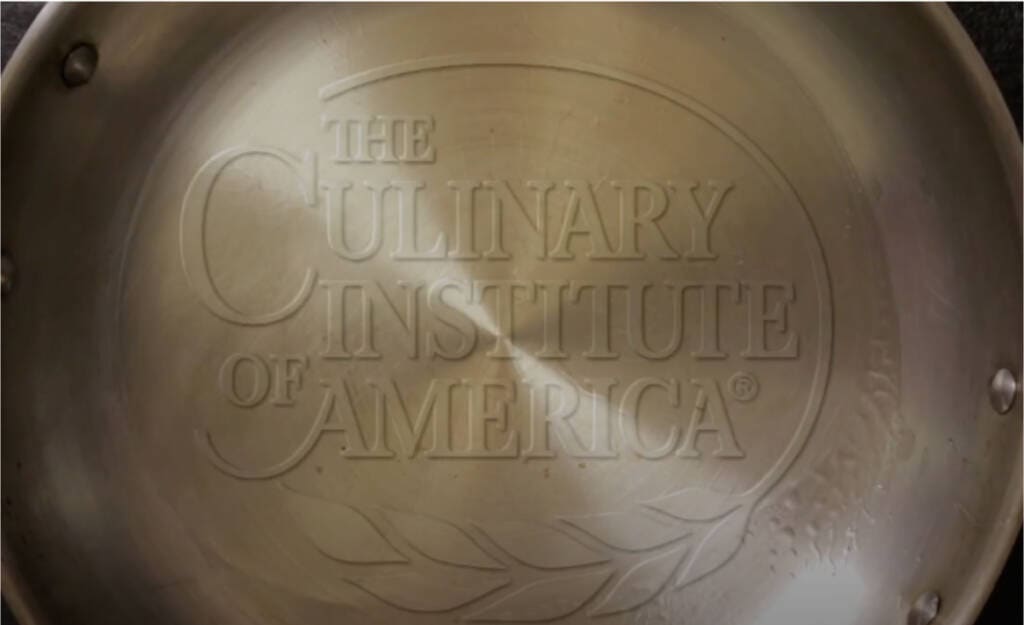 A metal pan with the embossed text “The Culinary Institute of America” in the center. The pan has a smooth surface with some light reflections and a few rivets visible near the top left and right edges.