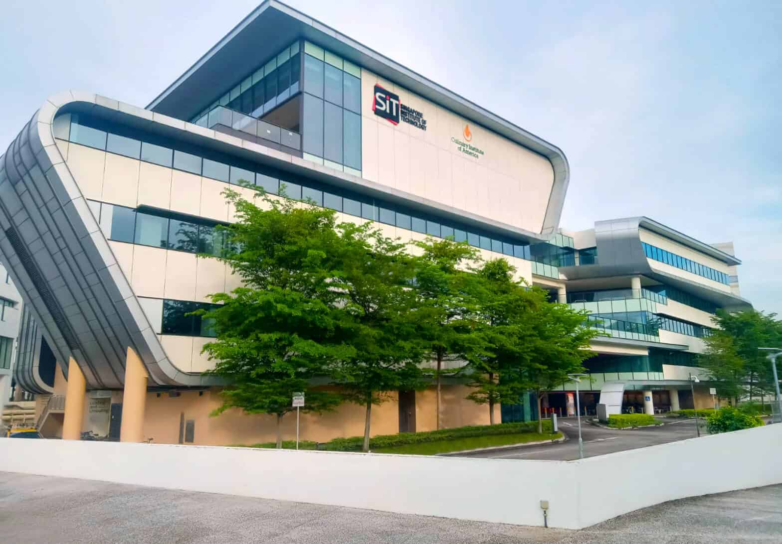 Modern multi-story building with large windows, featuring the "SIT" (Singapore Institute of Technology) and "Community" signs on the facade. It is surrounded by trees and greenery, with a clear blue sky above.